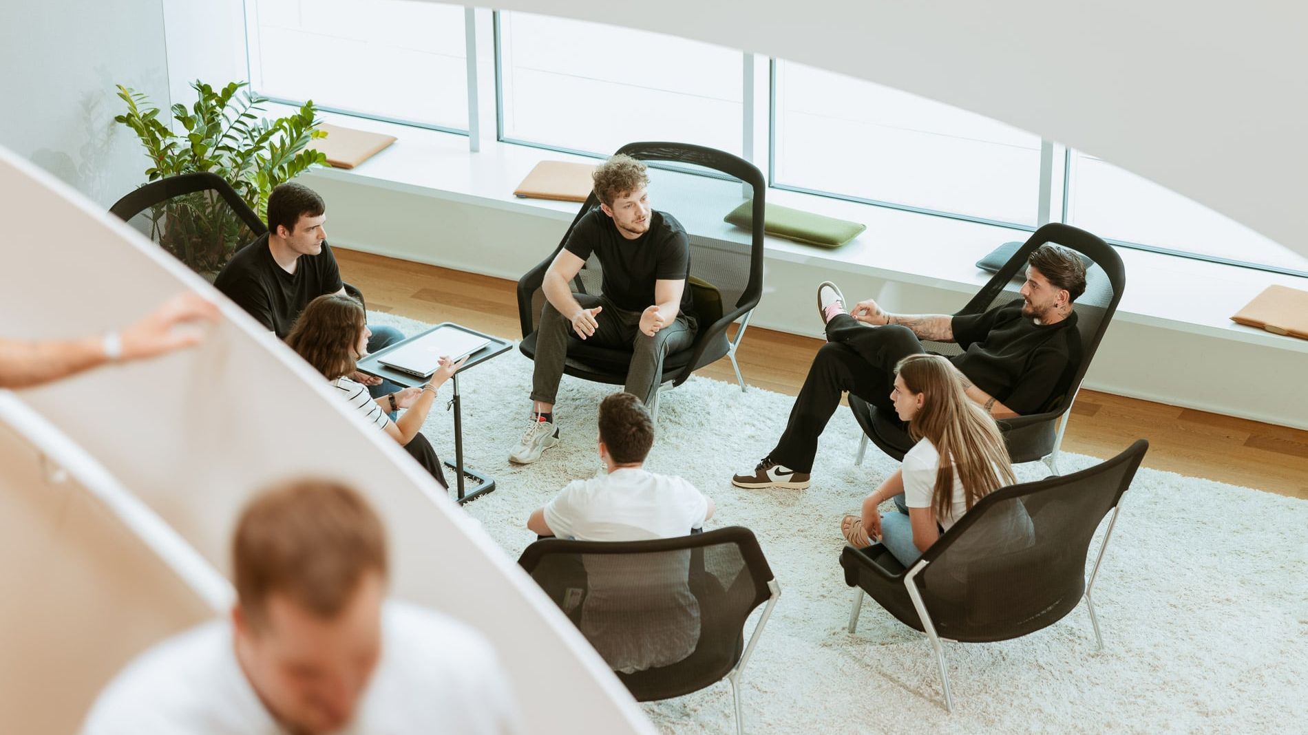 Gruppe von sechs Personen sitzt in einem modernen Büro auf bequemen Stühlen und unterhält sich. Sie sind in einem hellen Raum mit großen Fenstern und Pflanzen.