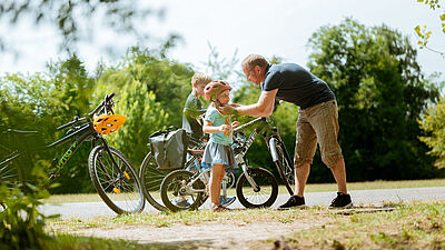 Ein Mann, wahrscheinlich der Papa, hilft einem kleinen Mädchen, das einen Fahrradhelm trägt, während ein Junge daneben steht. Im Hintergrund sind Fahrräder und grüne Bäume zu sehen.