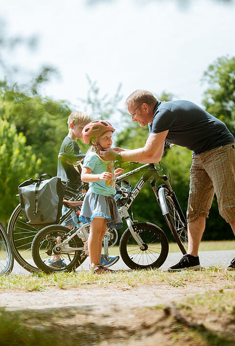 Ein Mann, wahrscheinlich der Papa, hilft einem kleinen Mädchen, das einen Fahrradhelm trägt, während ein Junge daneben steht. Im Hintergrund sind Fahrräder und grüne Bäume zu sehen.