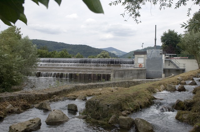 Das Bild zeigt das Wasserkraftwerk in Fahrnau. Es ist eine moderne Anlage mit mehreren Wasserfällen und einem Wasserkanal, der durch eine Betonstruktur fließt. Im Hintergrund sind Hügel und Bäume zu sehen.