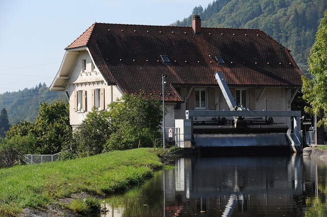 Das Bild zeigt das Wasserkraftwerk in Gündenhausen. Es ist ein historisches Gebäude mit einem roten Ziegeldach und einem Wasserkanal, der ruhig fließt. Im Hintergrund sind grüne Hügel und Bäume zu sehen.