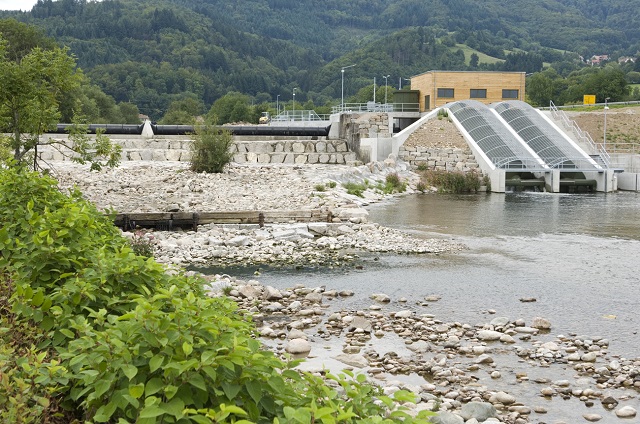 Bild zeigt das Wasserkraftwerk in Hausen Neumatt. Es ist eine moderne Anlage mit einem geneigten Gitter, das über einen Wasserkanal führt. Das Gebäude ist grau und von Bäumen und Felsen umgeben.
