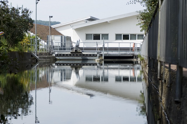 Das Bild zeigt das Wasserkraftwerk in Maulburg. Es ist eine moderne Anlage mit einem weißen Gebäude und einem Wasserkanal, der ruhig fließt. Im Hintergrund sind Bäume und ein Zaun zu sehen.