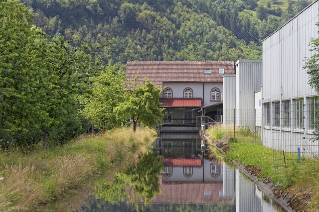 Das Bild zeigt das Wasserkraftwerk in Schappe. Es ist ein historisches Gebäude mit einem roten Ziegeldach und einem Wasserkanal, der ruhig fließt. Im Hintergrund sind grüne Hügel und Bäume zu sehen.