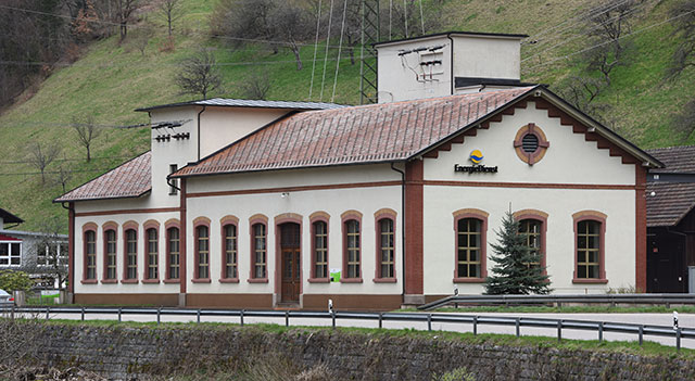 Das Bild zeigt das Wasserkraftwerk in Mambach. Es ist ein historisches Gebäude mit einem roten Ziegeldach und einem Wasserkanal, der ruhig fließt. Im Hintergrund sind grüne Hügel und Bäume zu sehen.