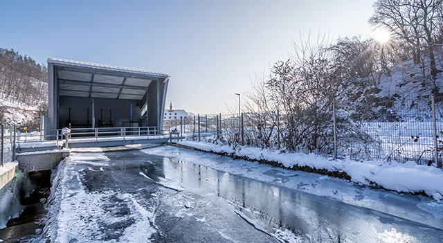 Das Bild zeigt das Wasserkraftwerk in Hausen, Bergwerk. Es ist eine moderne Anlage mit einem weißen Gebäude und einem Wasserkanal, der teilweise zugefroren ist. Im Hintergrund sind Bäume und Schnee zu sehen.