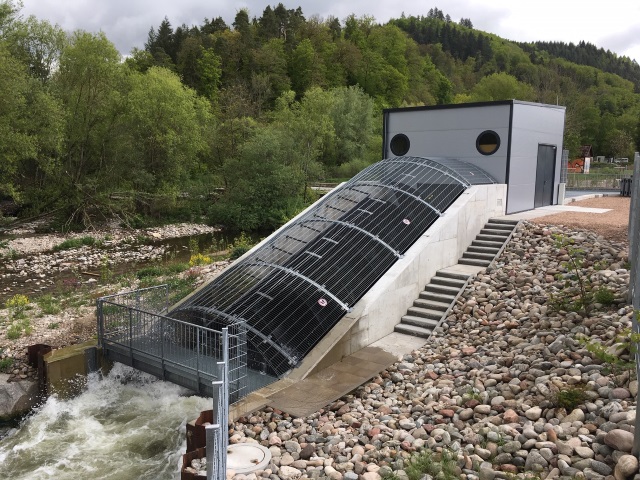 1.	Das Bild zeigt das Wasserkraftwerk in Maulburg. Es ist eine moderne Anlage mit einem geneigten Gitter, das über einen Wasserkanal führt. Das Gebäude ist grau und von Bäumen und Felsen umgeben.