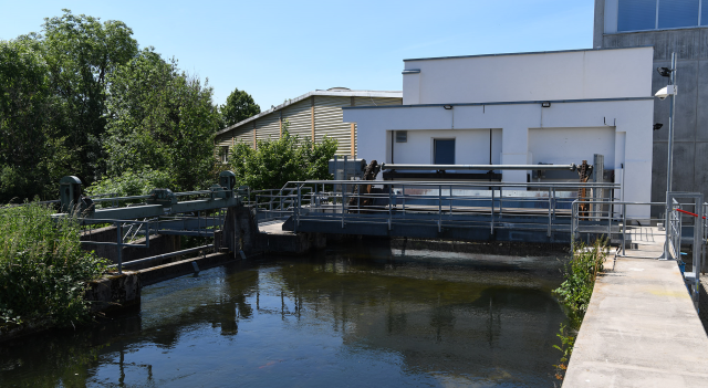 Das Bild zeigt das Wasserkraftwerk in Hausen Kummatt. Es ist eine moderne Anlage mit einem weißen Gebäude und einem Wasserkanal, der ruhig fließt. Im Hintergrund sind Bäume und ein Zaun zu sehen.