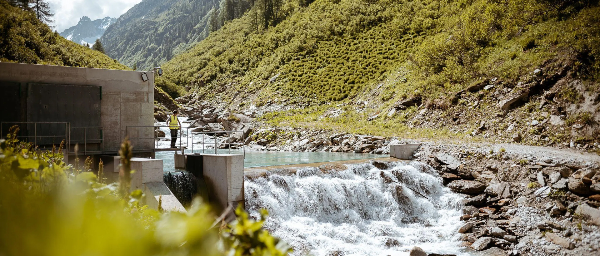 Ein Mitarbeiter von naturenergie in einer gelben Sicherheitsweste steht auf einer Plattform neben einem kleinen Wasserkraftwerk in einer bergigen Landschaft. Wasser fließt über eine Staumauer, umgeben von grüner Vegetation und felsigem Gelände